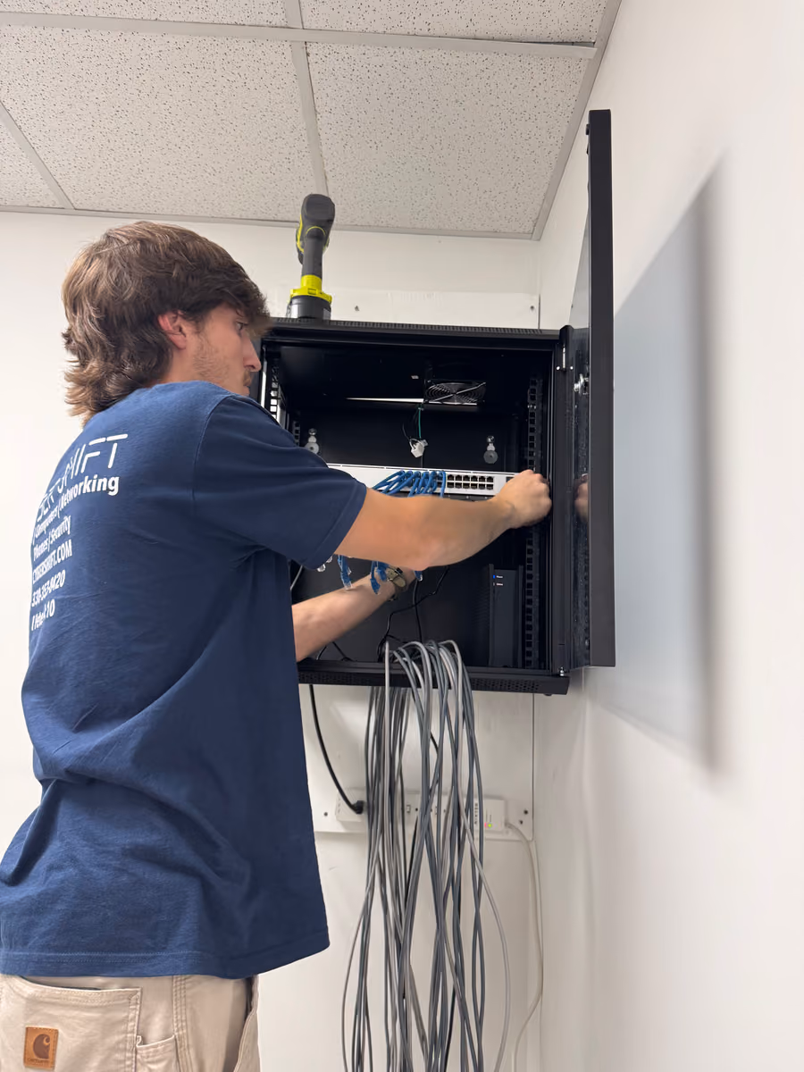 A Cyber Shift technician connecting blue patch cables to a switch in a wall-mounted network cabinet A Cyber Shift technician connecting blue patch cables to a switch in a wall-mounted network cabinet